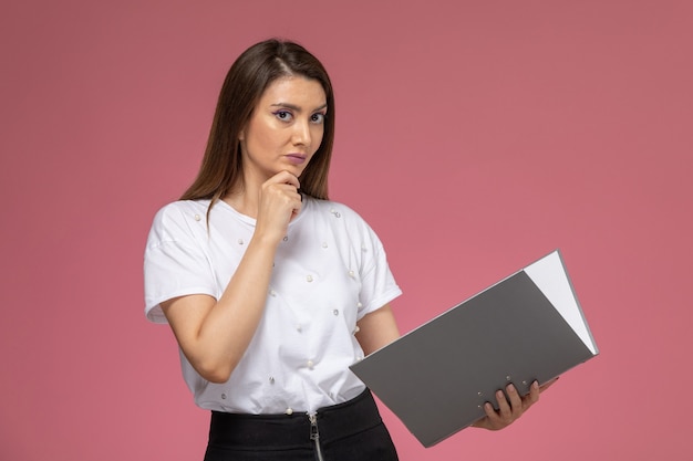 Vista frontal mujer joven en camisa blanca sosteniendo y leyendo un archivo gris en un escritorio rosa claro