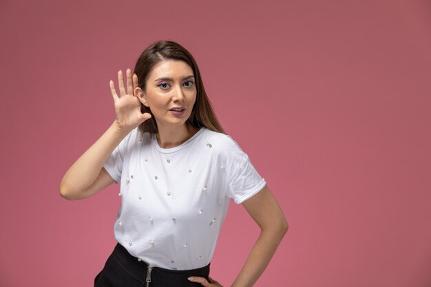 Vista frontal mujer joven con camisa blanca tratando de escuchar en la pared rosa, pose de mujer modelo