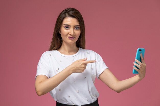 Vista frontal mujer joven con camisa blanca usando su teléfono en la pared rosa, mujer de color pose modelo mujer