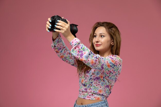 Vista frontal mujer joven en camisa de diseño floral y jeans sonriendo y sosteniendo la cámara de fotos en el fondo rosa