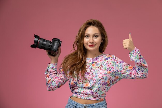 Vista frontal mujer joven en camisa de diseño floral y jeans sosteniendo cámara de fotos negra sobre fondo rosa