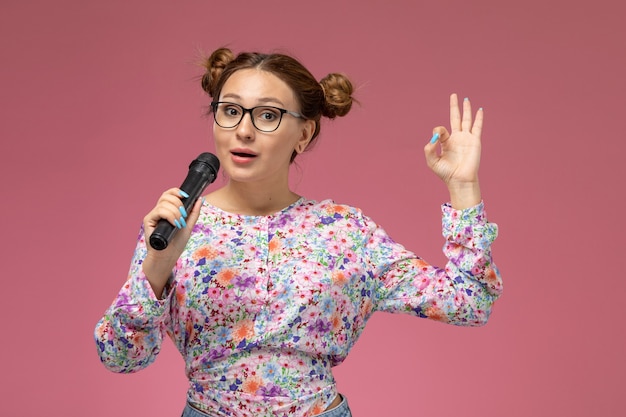 Vista frontal mujer joven en camisa de diseño floral y jeans sosteniendo el micrófono tratando de cantar en el modelado de fondo rosa