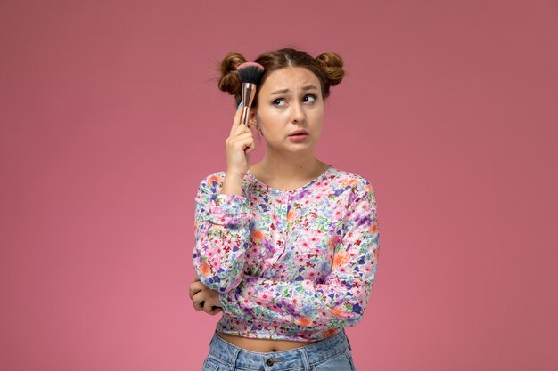 Vista frontal mujer joven en camisa de flor diseñada y jeans posando con pincel en la mano sobre el fondo rosa