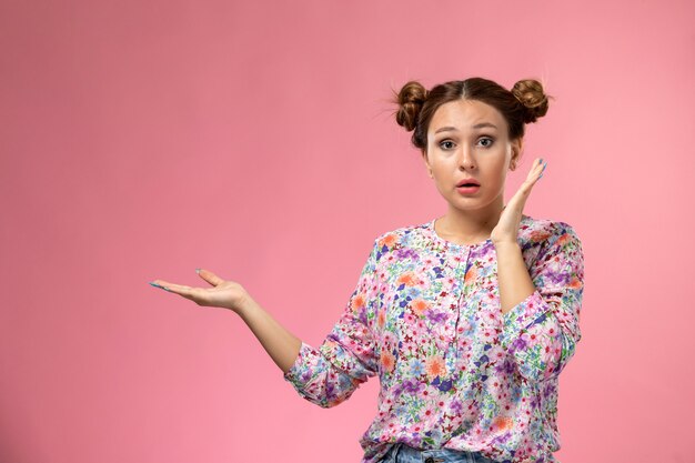 Vista frontal mujer joven en camisa de flor diseñada posando con expresión de sorpresa en el escritorio rosa