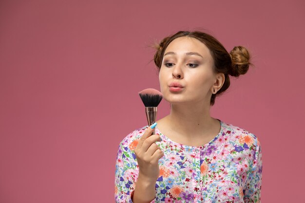 Vista frontal mujer joven en camisa de flor diseñada sosteniendo un pincel de maquillaje sobre el fondo rosa