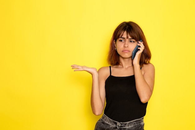Vista frontal de la mujer joven en camisa negra hablando por teléfono en la pared de color amarillo claro