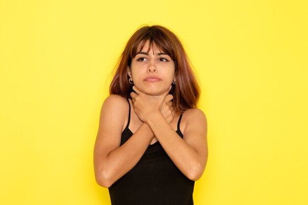 Vista frontal de la mujer joven en camisa negra y jeans grises ahogándose en la pared amarilla