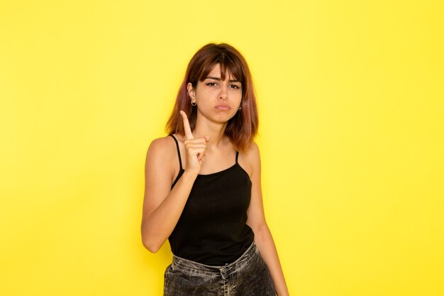 Vista frontal de la mujer joven en camisa negra y jeans grises amenazando con su dedo en la pared amarilla