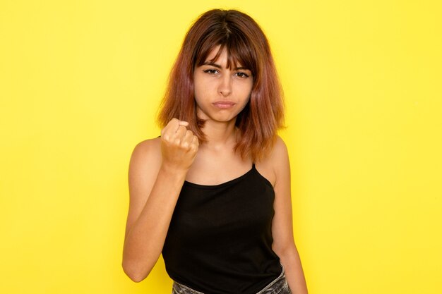 Vista frontal de la mujer joven en camisa negra y jeans grises amenazando con su puño en la pared amarilla