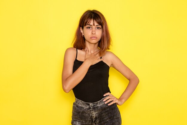 Vista frontal de la mujer joven en camisa negra y jeans grises posando en la pared amarilla