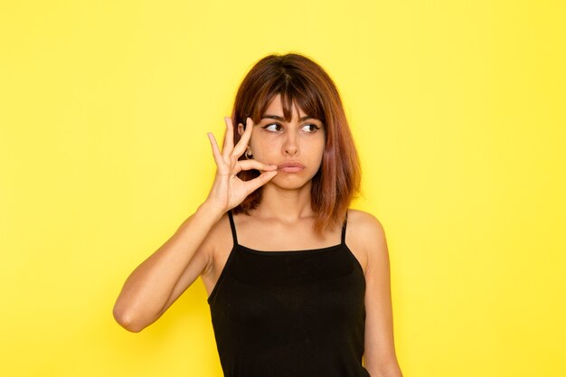 Vista frontal de la mujer joven en camisa negra y jeans grises posando en la pared amarilla