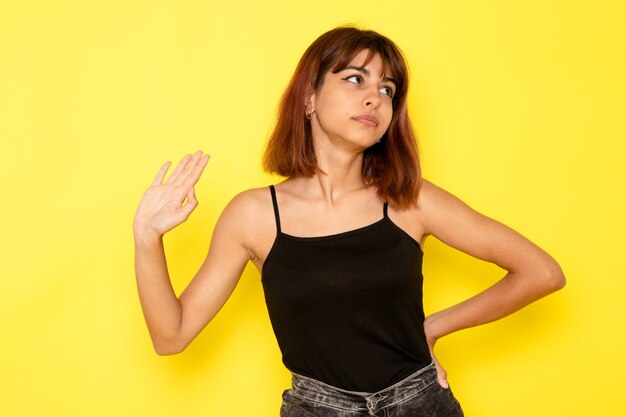 Vista frontal de la mujer joven en camisa negra y jeans grises simplemente posando en la pared amarilla