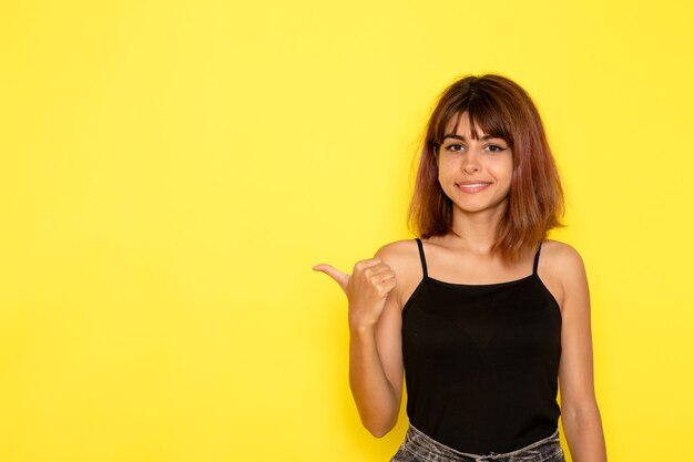 Vista frontal de la mujer joven en camisa negra y jeans grises sonriendo y posando en la pared amarilla