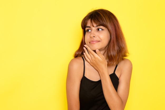 Vista frontal de la mujer joven en camisa negra y jeans grises sonriendo y posando en la pared amarilla