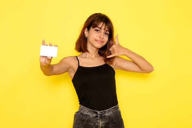 Vista frontal de la mujer joven en camisa negra y jeans grises con tarjeta blanca en la pared amarilla