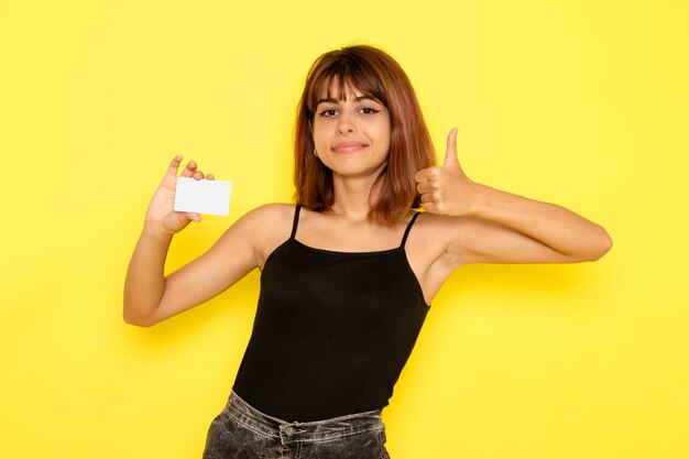 Vista frontal de la mujer joven en camisa negra y jeans grises con tarjeta en la pared amarilla