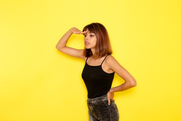 Vista frontal de la mujer joven en camisa negra mirando a lo lejos en la pared de color amarillo claro