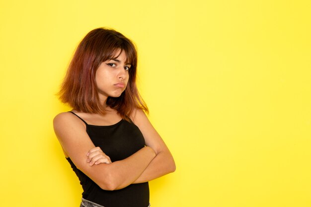 Vista frontal de la mujer joven en camisa negra posando con expresión enojada en la pared de color amarillo claro