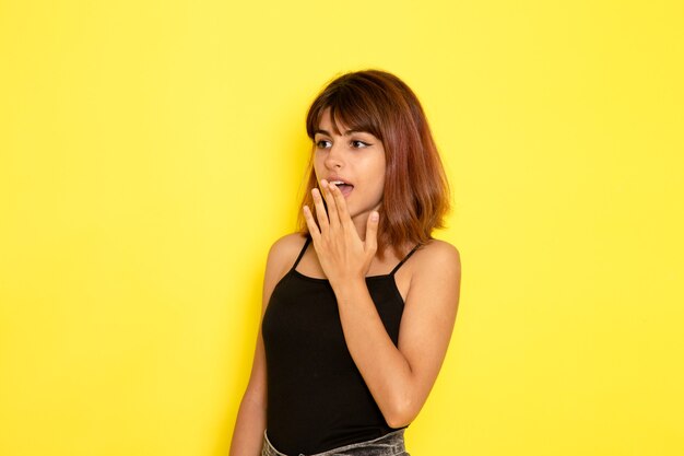 Vista frontal de la mujer joven en camisa negra posando en la pared de color amarillo claro