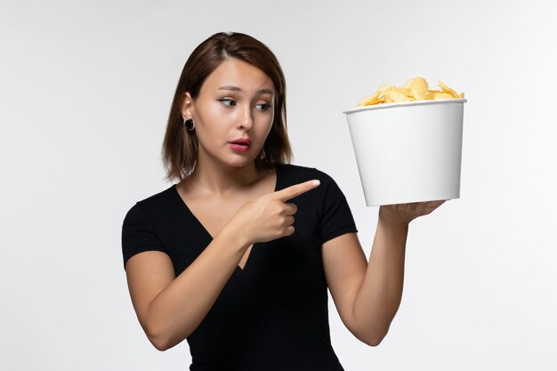 Vista frontal mujer joven en camisa negra sosteniendo papas fritas y posando en el escritorio blanco