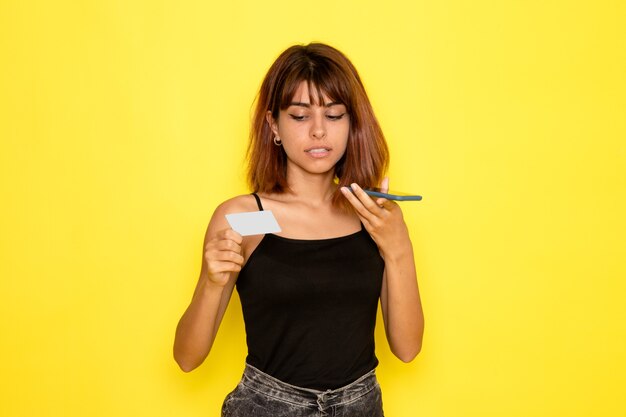 Vista frontal de la mujer joven en camisa negra sosteniendo la tarjeta y hablando por teléfono en la pared de color amarillo claro