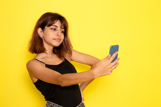 Vista frontal de la mujer joven en camisa negra tomando un selfie en la pared de color amarillo claro