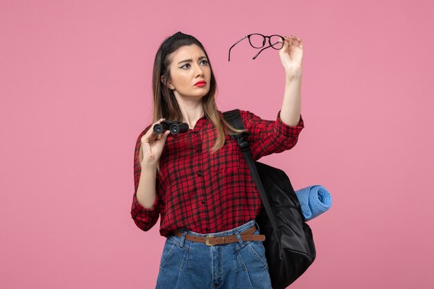 Vista frontal mujer joven en camisa roja con binoculares en el modelo de color de mujer de fondo rosa