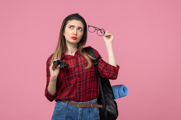 Vista frontal mujer joven en camisa roja con binoculares en el modelo de color de mujer de fondo rosa