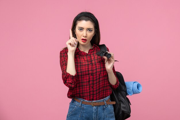 Vista frontal mujer joven en camisa roja con binoculares en la mujer de color de estudiante de fondo rosa