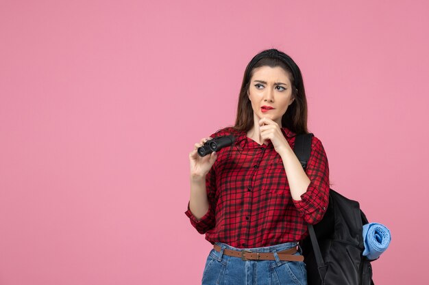 Vista frontal mujer joven en camisa roja con binoculares en una mujer de color rosa estudiante de escritorio