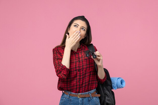 Vista frontal mujer joven en camisa roja con binoculares sobre fondo rosa claro color de moda mujer