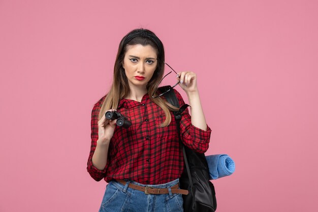 Vista frontal mujer joven en camisa roja con binoculares sobre fondo rosa claro modelo de color de mujer