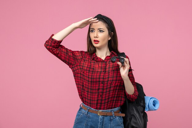 Vista frontal mujer joven en camisa roja con binoculares sobre fondo rosa color estudiante mujer