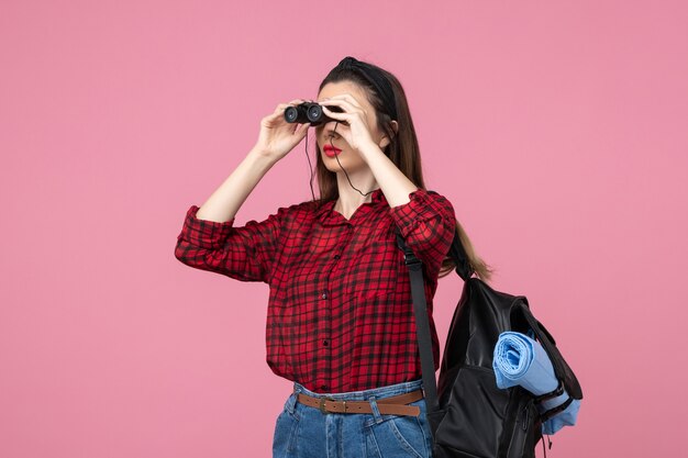 Vista frontal mujer joven en camisa roja con binoculares sobre fondo rosa mujer de color de estudiante