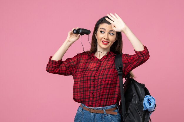 Vista frontal mujer joven en camisa roja con binoculares sobre fondo rosa mujer de colores de estudiante