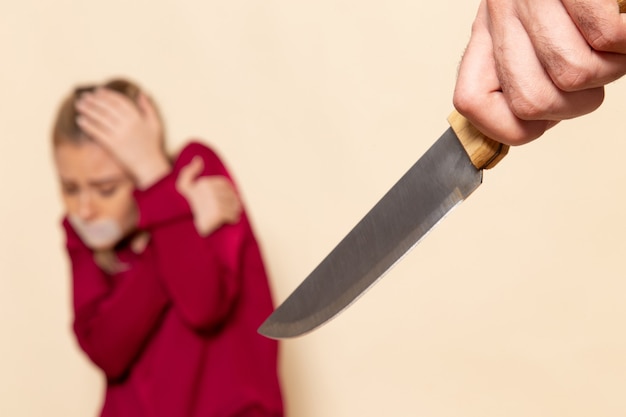 Foto gratuita vista frontal mujer joven en camisa roja con la boca atada asustada del cuchillo en el espacio de la crema tela femenina