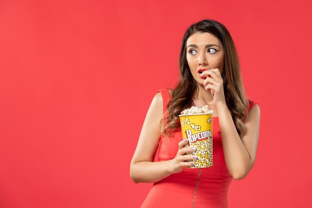 Vista frontal mujer joven en camisa roja comiendo palomitas de maíz viendo la película en la superficie roja