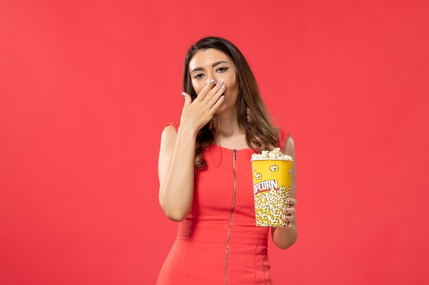 Vista frontal mujer joven en camisa roja sosteniendo palomitas de maíz y bostezando en la superficie de color rojo claro