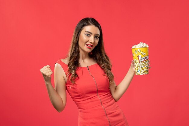 Vista frontal mujer joven en camisa roja sosteniendo palomitas de maíz en la superficie de color rojo claro