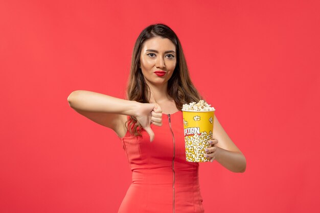 Vista frontal mujer joven en camisa roja sosteniendo palomitas de maíz en la superficie de color rojo claro