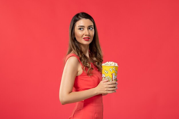 Vista frontal mujer joven en camisa roja sosteniendo palomitas de maíz viendo películas en la superficie roja