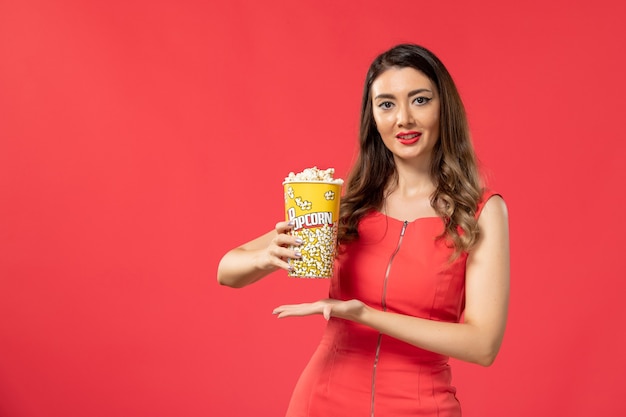 Vista frontal mujer joven en camisa roja sosteniendo el paquete de palomitas de maíz y sonriendo sobre la superficie roja