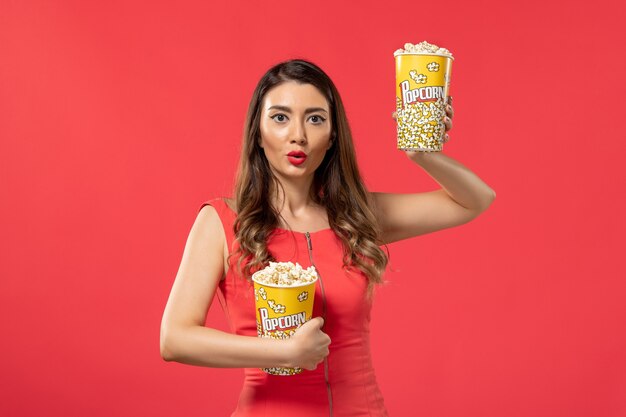 Vista frontal mujer joven en camisa roja sosteniendo paquetes de palomitas de maíz en la superficie roja