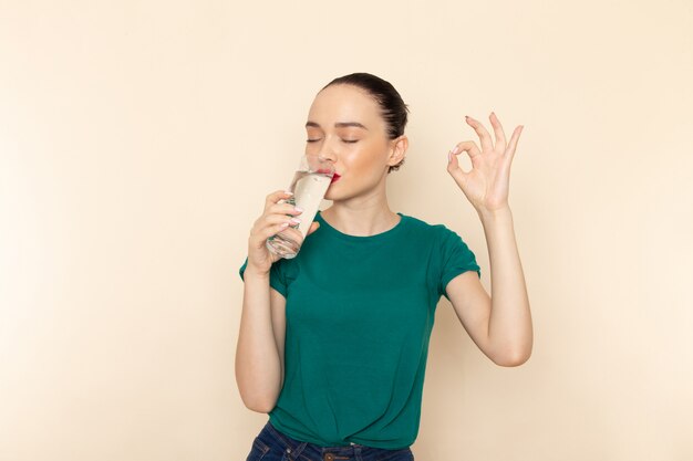 Vista frontal mujer joven en camisa verde oscuro y jeans bebiendo vaso de agua en beige