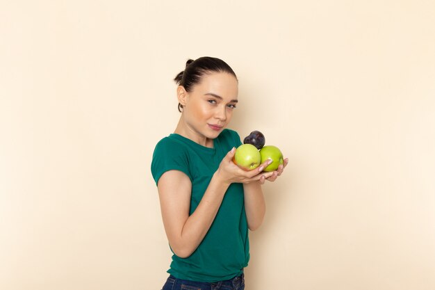 Vista frontal mujer joven en camisa verde oscuro y jeans con frutas en beige