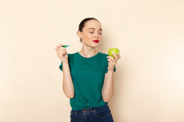 Vista frontal mujer joven en camisa verde oscuro y jeans con manzana e inyección en beige