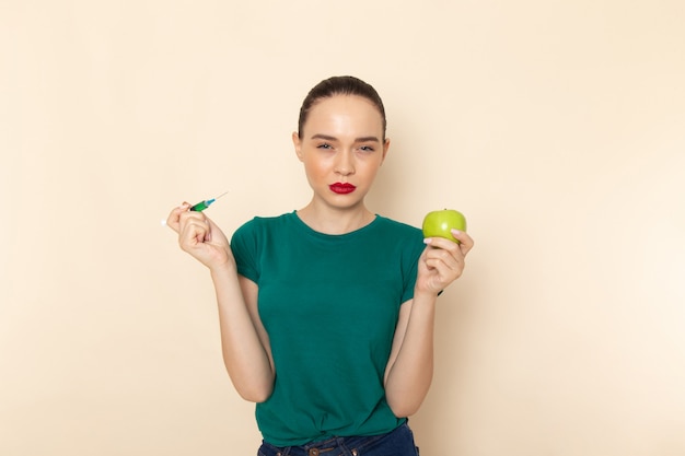 Vista frontal mujer joven en camisa verde oscuro y jeans con manzana e inyección en beiges