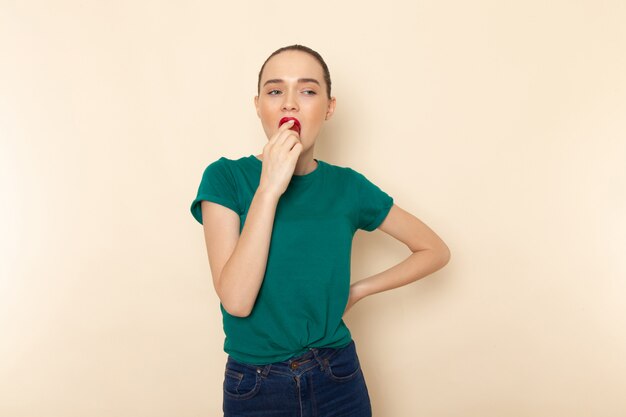 Vista frontal mujer joven en camisa verde oscuro y jeans mordiendo fruta en beige