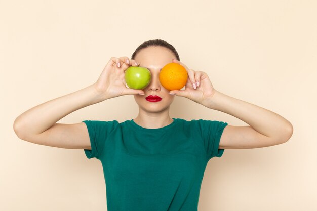 Vista frontal mujer joven en camisa verde oscuro y jeans con naranja y manzana cubriéndose los ojos en beige