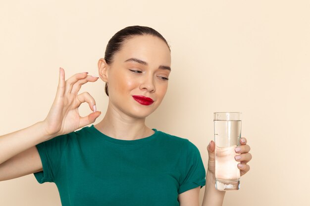 Vista frontal mujer joven en camisa verde oscuro y jeans sosteniendo un vaso de agua sonriendo en beige